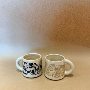 Può includere: Due tazze in ceramica con manici spessi e arrotondati. La tazza a sinistra presenta un motivo astratto marmorizzato in bianco e nero, mentre quella a destra mostra un design grafico lineare color sabbia e bianco. Entrambe hanno una finitura interna liscia.