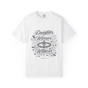 以下が含まれることがあります： 黒いグラフィックプリントが施された白い半袖Tシャツ。デザインには「Daughter」「Warrior」「Witness」という言葉、茨の冠に囲まれた剣、そして花の要素が含まれています。グラフィックの下には「1 Peter 2:9」というテキストが印刷されています。