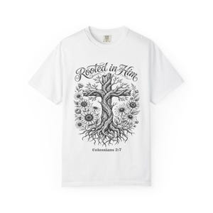 May include: A white short-sleeve t-shirt featuring a black ink-style graphic of a cross formed by tree roots and branches, surrounded by sunflowers. The text "Rooted in Him" appears above the cross, and "Colossians 2:7" is printed below it.