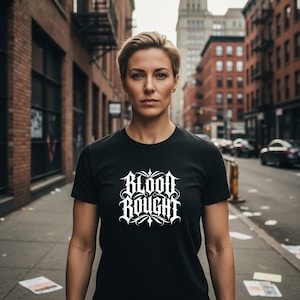 May include: A black short-sleeved t-shirt featuring a white graphic print on the chest with the text "BLOOD BOUGHT" in a stylized, decorative font. The shirt has a crew neckline and a standard fit, constructed from a solid black fabric.