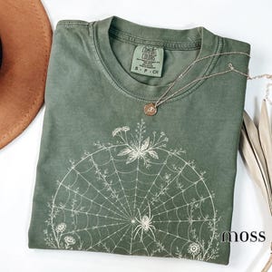 May include: A moss green short-sleeve t-shirt featuring a white graphic print of a spider web adorned with flowers and a spider. The shirt has a crew neckline and a Comfort Colors brand label inside the collar. A thin gold chain necklace rests on top.