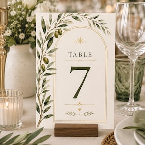 May include: A rectangular table number card featuring the text TABLE 7 in dark green font. The card is decorated with a watercolor illustration of olive branches and green olives along the left side and top, framed by a thin gold arch and small heart icons.