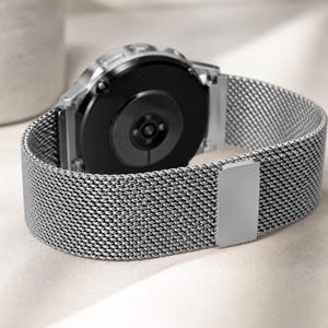 May include: A silver-toned smartwatch featuring a circular black case back and a flexible stainless steel mesh strap. The watch band includes a rectangular metal sliding clasp mechanism for secure adjustment around the wrist.