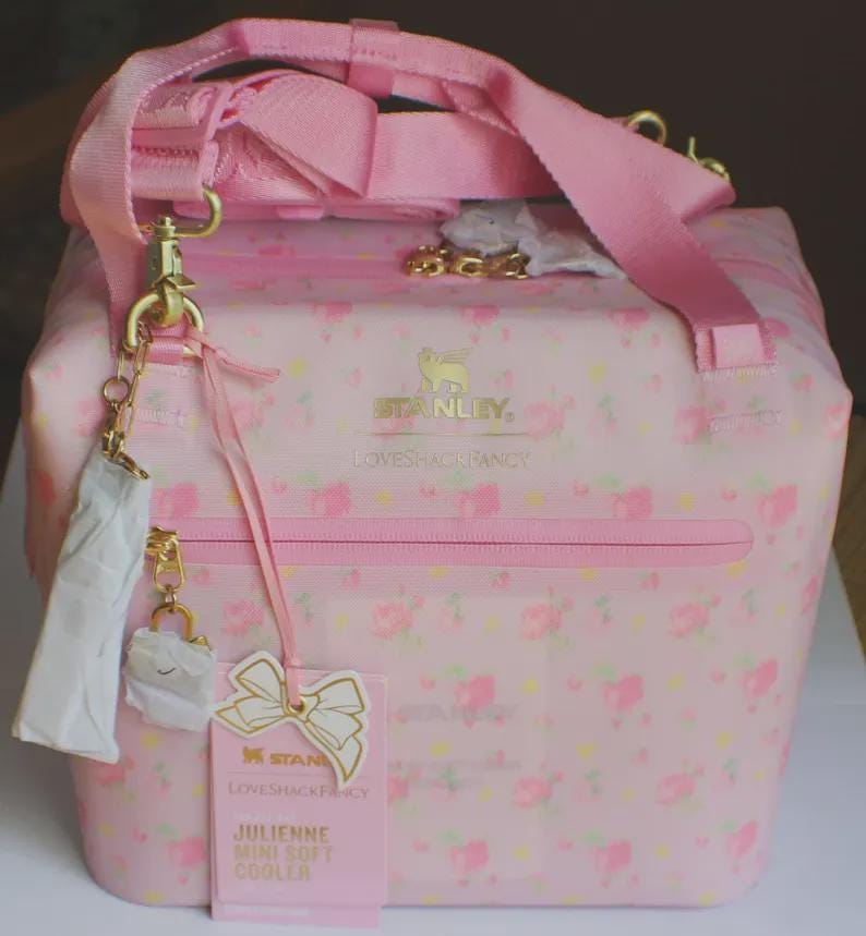 May include: A light pink soft cooler bag featuring a floral pattern, gold hardware, and a pink adjustable shoulder strap. The front displays the Stanley and LoveShackFancy logos, a zippered pocket, and a product tag labelled Julienne Mini Soft Cooler.