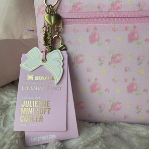 May include: A light pink soft cooler with a floral pattern, featuring a gold-tone heart charm and a bow-shaped tag. The tag displays the text Stanley LoveShackFancy The All Day Julienne Mini Soft Cooler 7.4 QT | 7.0 L and Stanley1913.com.