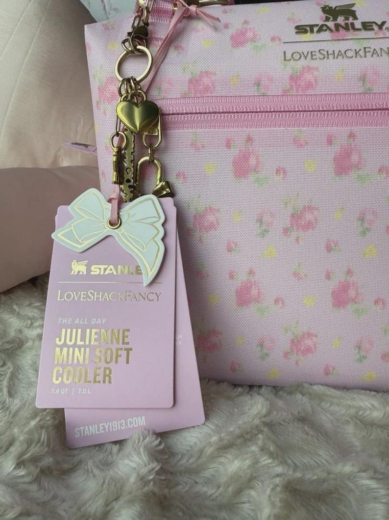 May include: A light pink soft cooler with a floral pattern, featuring a gold-tone heart charm and a bow-shaped tag. The tag displays the text Stanley LoveShackFancy The All Day Julienne Mini Soft Cooler 7.4 QT | 7.0 L and Stanley1913.com.