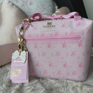 May include: A light pink soft cooler bag featuring a floral pattern, gold-tone hardware, and a zippered front pocket. The bag displays the Stanley and LoveShackFancy logos, includes a gold heart charm, and has a product tag labelled Julienne Mini Soft Cooler.