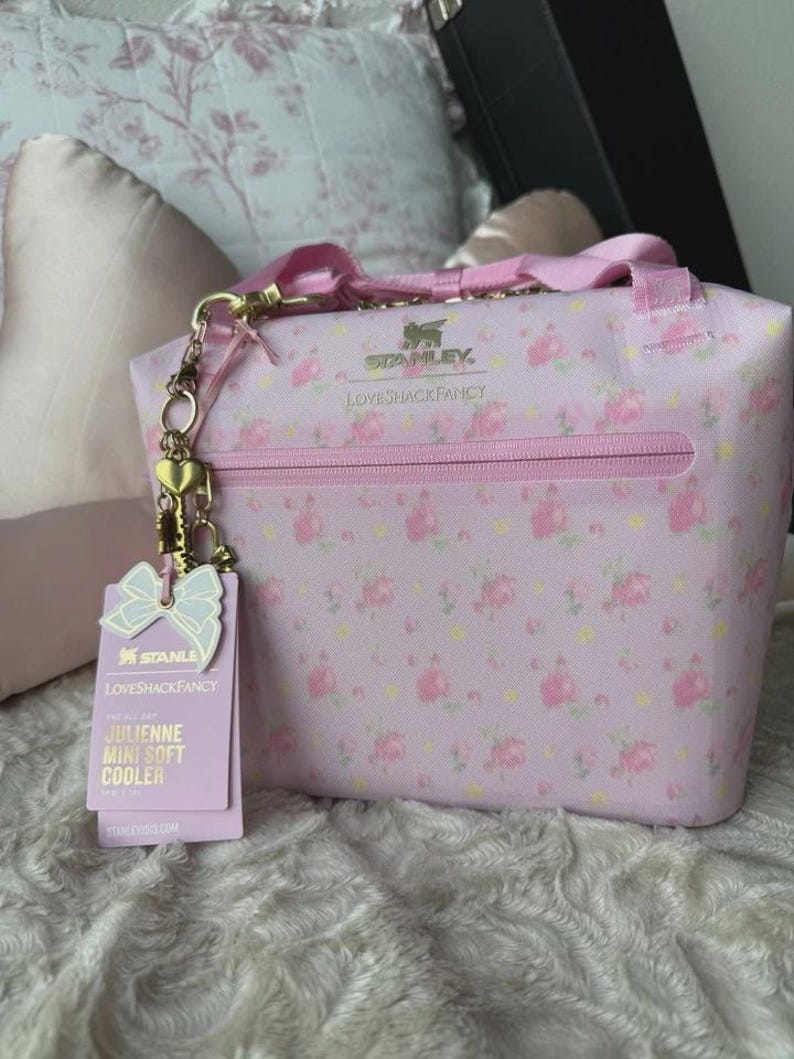 May include: A light pink soft cooler bag featuring a floral pattern, gold-tone hardware, and a zippered front pocket. The bag displays the Stanley and LoveShackFancy logos, includes a gold heart charm, and has a product tag labelled Julienne Mini Soft Cooler.