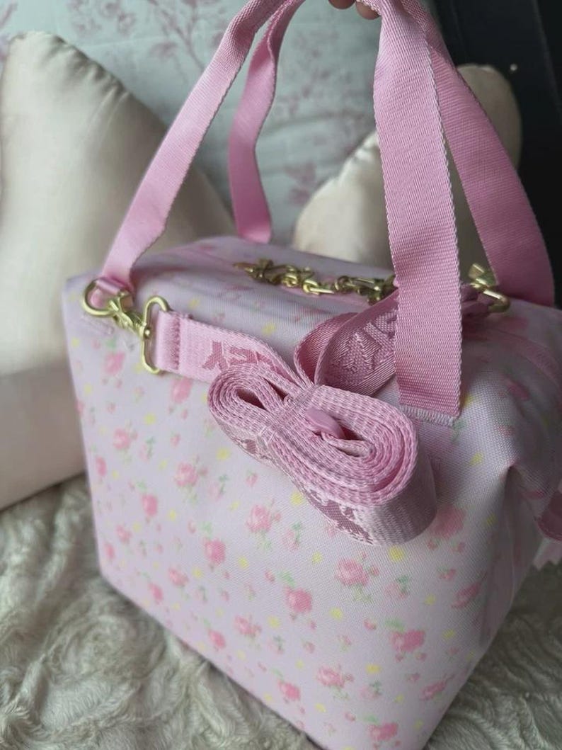 May include: A light pink rectangular bag featuring a repeating floral pattern of small pink roses and yellow dots. The bag includes two pink fabric top handles and a detachable, adjustable pink shoulder strap with gold-toned metal hardware clips.