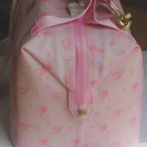 May include: A light pink duffel bag featuring a repeating pattern of small pink roses and yellow dots. The bag includes a vertical pink zip closure, pink fabric handles, and a gold-toned metal clip. A small gold logo is visible on the zip tab.