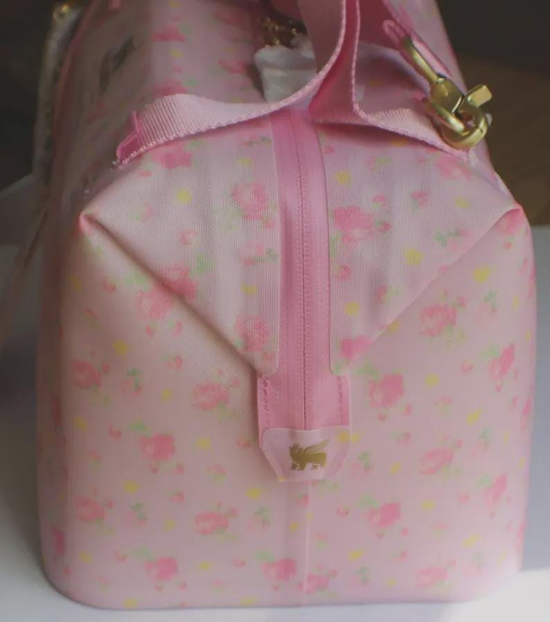 May include: A light pink duffel bag featuring a repeating pattern of small pink roses and yellow dots. The bag includes a vertical pink zip closure, pink fabric handles, and a gold-toned metal clip. A small gold logo is visible on the zip tab.