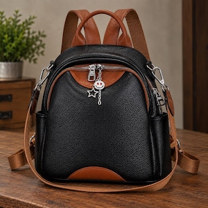 Puede incluir: Una mochila de cuero negro granulado con detalles en cuero color canela, que cuenta con un asa superior, correas ajustables del mismo tono y herrajes plateados. El tirador de la cremallera frontal tiene un dije de estrella y una cara sonriente.