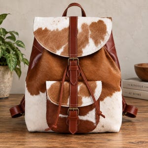 May include: A backpack featuring a brown and white cowhide pattern with smooth brown leather accents. The bag includes a large main flap, a smaller front pocket, and vertical brown leather straps with metal buckles for secure closure.