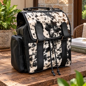 Puede incluir: Una mochila con estampado de piel de vaca en blanco y negro en la solapa frontal y el cuerpo, acentuada con ribetes de cuero sintético negro, dos correas verticales con hebillas, un asa superior y un bolsillo lateral con cremallera.