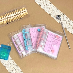May include: A dental study model with teeth, a metal dental mirror, a blue dental floss container, and three small clear plastic binders with pink inserts featuring text and ring mechanisms in pastel pink, blue, and purple colours.