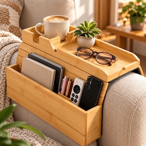 May include: A bamboo sofa armrest organizer featuring a flat top surface for a mug, a small potted plant, and eyeglasses. The front section includes a deep pocket for notebooks, a small compartment for pens, a remote control, and a smartphone.