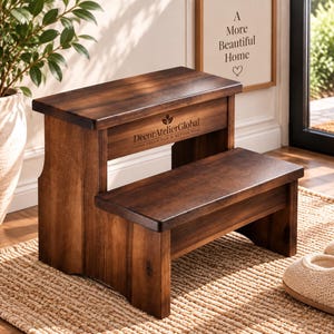 May include: A two-step wooden stool with a dark brown finish. The front panel features the text DecorAtelierGlobal and the phrase HOME DECOR FOR A BETTER YOU. The stool has a rectangular top step and a lower step, both with smooth surfaces.