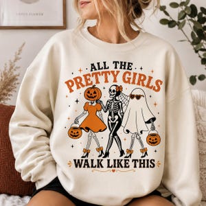 May include: A cream-coloured crewneck sweatshirt featuring a graphic print with the text ALL THE PRETTY GIRLS WALK LIKE THIS. The design shows three figures: a pumpkin-headed character in a dress, a skeleton, and a ghost wearing heart-shaped sunglasses.