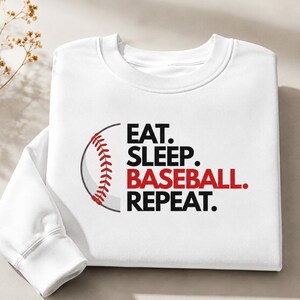 May include: A white crewneck sweatshirt featuring a graphic of a baseball with red stitching on the left side. To the right of the baseball, the text reads EAT. SLEEP. BASEBALL. REPEAT. in black and red lettering, with BASEBALL in red.