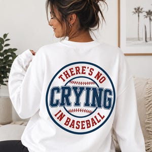 Op de afbeelding: Een wit sweatshirt met lange mouwen en een ronde hals, voorzien van een grote cirkelvormige afbeelding op de achterkant. Het ontwerp bevat een honkbalillustratie met de rode en blauwe tekst "THERE'S NO CRYING IN BASEBALL" in een cirkel.