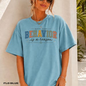 Könnte beinhalten: Ein hellblaues T-Shirt mit kurzen Ärmeln und dem Text BEHIND EVERY BEHAVIOR is a reason in mehrfarbiger Block- und Schreibschrift. Das Design enthält ein kleines Herzsymbol über dem Text und ein Puzzleteil-Symbol unter dem Text.