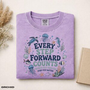 Könnte beinhalten: Ein hellviolettes T-Shirt mit einer Grafik von Meerestieren wie einer Schildkröte, Quallen und Fischen. Der Text lautet: EVERY STEP FORWARD COUNTS, PROGRESS OVER PERFECTION, EVERY STEP MATTERS. Das T-Shirt hat ein Comfort Colors Markenetikett.