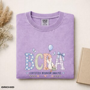 Könnte beinhalten: Ein hellviolettes T-Shirt mit dem Text BCBA CERTIFIED BEHAVIOR ANALYST. Das Design zeigt eine weiße Ente mit einem blauen Luftballon, Schmetterlingen und einer blauen Schleife. Die Buchstaben haben Blumen-, Streifen- und Vichy-Karomuster.