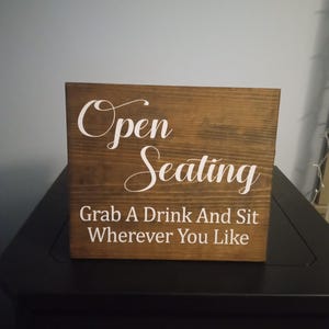 May include: A rectangular wooden sign with a dark brown stained finish and white lettering. The text reads Open Seating in a script font, followed by the text Grab A Drink And Sit Wherever You Like in a simple, clean, sans-serif typeface.