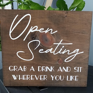 May include: A rectangular dark brown wooden sign with a visible wood grain and a knot. The sign features white text that reads Open Seating in a script font, followed by the text GRAB A DRINK AND SIT WHEREVER YOU LIKE in a sans-serif font.
