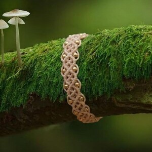 Peut inclure: Un bracelet en macramé beige présentant un motif de nœuds en forme de losange répété, intégré avec de petites perles métalliques dorées polies. Le bracelet est présenté sur une branche d'arbre recouverte de mousse près de deux petits champignons blancs.