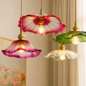 May include: Four pendant lights with flower-shaped glass shades in red, pink, green, and clear, each featuring a brass-toned fixture and a warm glowing light bulb. The shades have a fluted, petal-like texture and hang from twisted fabric cords.