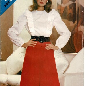 May include: A sewing pattern envelope labeled See & Sew 5084 Size A (8-10-12) featuring a white long-sleeved blouse with vertical ruffles on the chest, paired with a red knee-length skirt and a wide black cutout belt with a rectangular buckle.