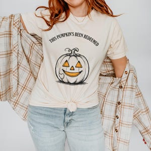 May include: A cream-colored short-sleeve t-shirt featuring a black line-art illustration of a jack-o'-lantern with orange-colored eyes and mouth. The text "THIS PUMPKIN'S BEEN REDEEMED" is printed in black capital letters above the pumpkin graphic.