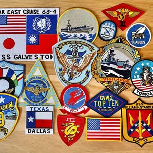 Pode incluir: Uma coleção de emblemas militares bordados com várias formas e designs, incluindo insígnias do USS Galveston, da Sétima Frota dos EUA, Seabees, Tomcat e Texas Dallas, juntamente com bandeiras americanas e diversos emblemas navais.