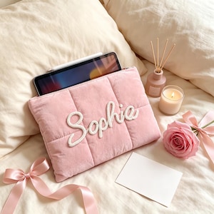 Könnte beinhalten: Eine gesteppte, hellrosa Tablethülle aus Samt mit dem Namen Sophie in weißer Schreibschrift auf der Vorderseite. Die Hülle enthält ein Tablet mit einem darauf liegenden Eingabestift und ist mit einem rosa Band und einer Rose verziert.