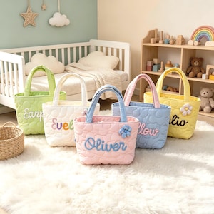 May include: A collection of five quilted tote bags in light green, white, light blue, pink, and yellow. Each bag features a unique name embroidered in cursive script on the front, along with a small, matching fabric flower accent attached.