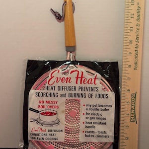 May include: A round metal heat diffuser with a light wood handle and a hanging loop, packaged in plastic. The label reads Even Heat, Heat Diffuser Prevents Scorching and Burning of Foods. A wooden ruler shows the tool is approximately 15 inches long.