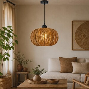 Peut inclure: Une suspension en forme de citrouille dotée d'un abat-jour en corde tressée beige, suspendue par un cordon et une rosace noirs. La lumière diffuse un éclat chaud à travers la texture de la fibre naturelle, formant une silhouette arrondie.