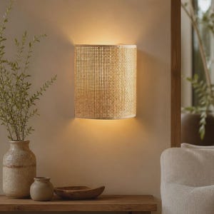 May include: A semi-cylindrical wall sconce featuring a light tan woven rattan shade. The fixture emits a warm, soft glow from both the top and bottom, casting light onto the wall surface. The design is simple, curved, and textured.