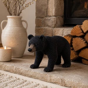 May include: A handcrafted crochet black bear figurine featuring a textured dark yarn body, rounded ears, and a light brown muzzle. The bear is depicted in a standing position with visible stitched details on its paws and a small black nose.