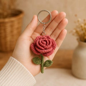 Può includere: Un portachiavi all'uncinetto a forma di rosa rosa scuro con un gambo verde e due piccole foglie verdi. Il fiore è fissato a un anello metallico color argento tramite una catenella. L'oggetto presenta punti di lavorazione a maglia visibili.