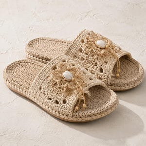 May include: A pair of beige crochet slide sandals featuring an open-toe design with a textured woven pattern. Each sandal is adorned with a central crocheted flower detail, a white oval shell accent, and small wooden beads on dangling strings.