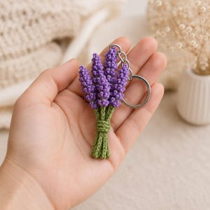 Può includere: Un portachiavi all'uncinetto a forma di piccolo bouquet di quattro rametti di lavanda viola con capolini testurizzati e annodati, e steli verdi legati insieme con un filo verde, fissato a un anello in metallo argentato.