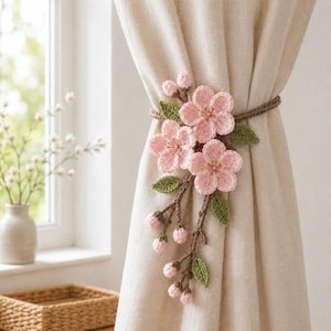 May include: A crochet curtain tieback featuring three light pink cherry blossom flowers, several small closed buds, and green leaves attached to a brown branch-like stem, wrapped around a plain beige fabric curtain.