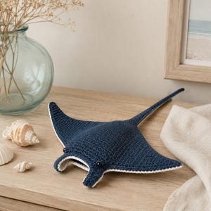 May include: A dark blue crocheted stingray plush toy with a white underside border and two small black bead eyes. The stingray features a flat, wide body shape with pointed wings and a long, slender tail extending from the centre of the back.