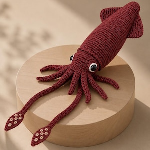 May include: A dark red crocheted squid toy featuring a textured yarn body, two long tentacles with light-colored circular suction cup details at the ends, and two black and white plastic eyes positioned on the head of the marine creature.