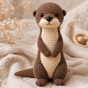 May include: A crocheted otter soft toy featuring dark brown yarn for the head, back, and tail, with a contrasting light beige yarn on the chest and face. The otter has small black safety eyes, a black nose, and thin white whiskers.