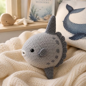 May include: A gray crochet sunfish plush toy with a round body, a small dorsal fin, a ventral fin, and a textured back edge. The fish features a small white mouth and a black bead eye. The surface of the toy is crafted from yarn stitches.