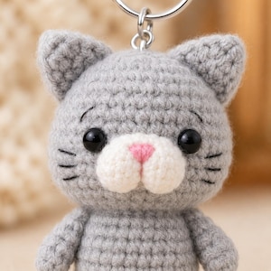 May include: A gray crocheted cat keychain featuring a round head with pointed ears, black plastic eyes, a white muzzle, and a small pink stitched nose. The cat has black embroidered whiskers on both cheeks and is attached to a silver metal ring.
