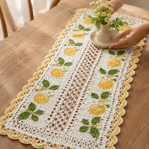 May include: A rectangular white crochet table runner featuring a repeating pattern of yellow citrus fruits, green leaves, and small white flowers. The runner has a scalloped yellow and white border and an intricate openwork lace design throughout.
